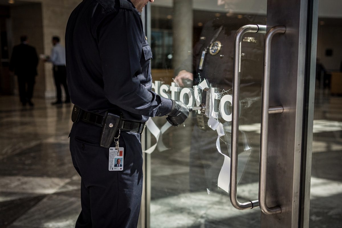 Harsh afternoon sunlight cutting across a glass-fronted corporate lobby, a security guard seen from the chest down using a metal scraper to peel white vinyl.