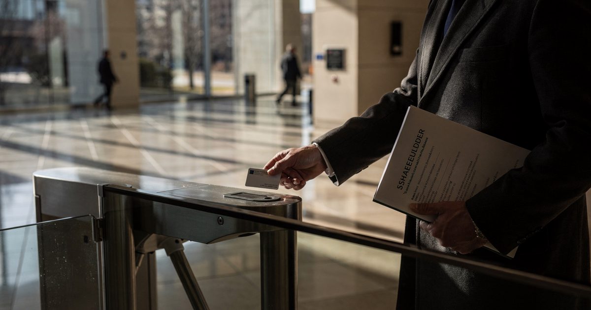 Harsh late-day sun cutting across a glass office lobby in Omaha, a person cropped at the shoulders in a charcoal coat feeding a keycard into a waist-high.