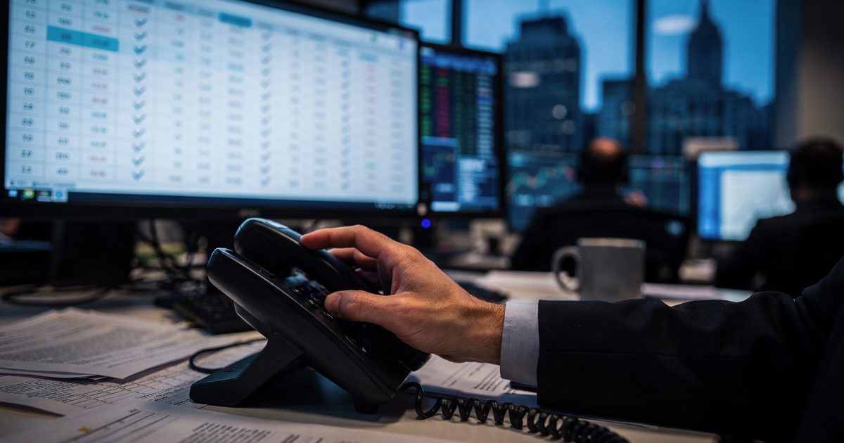 Close detail in the blue glow of Sunday dusk on a Manhattan hedge fund trading desk, an analyst's hand seen from the wrist down gripping a desk phone receiver.