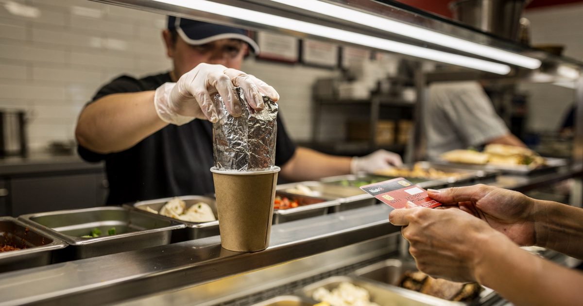 Medium shot from the customer side of a Chipotle service counter at harsh noon under white fluorescent strips, a crew member cropped below the visor pressing a.