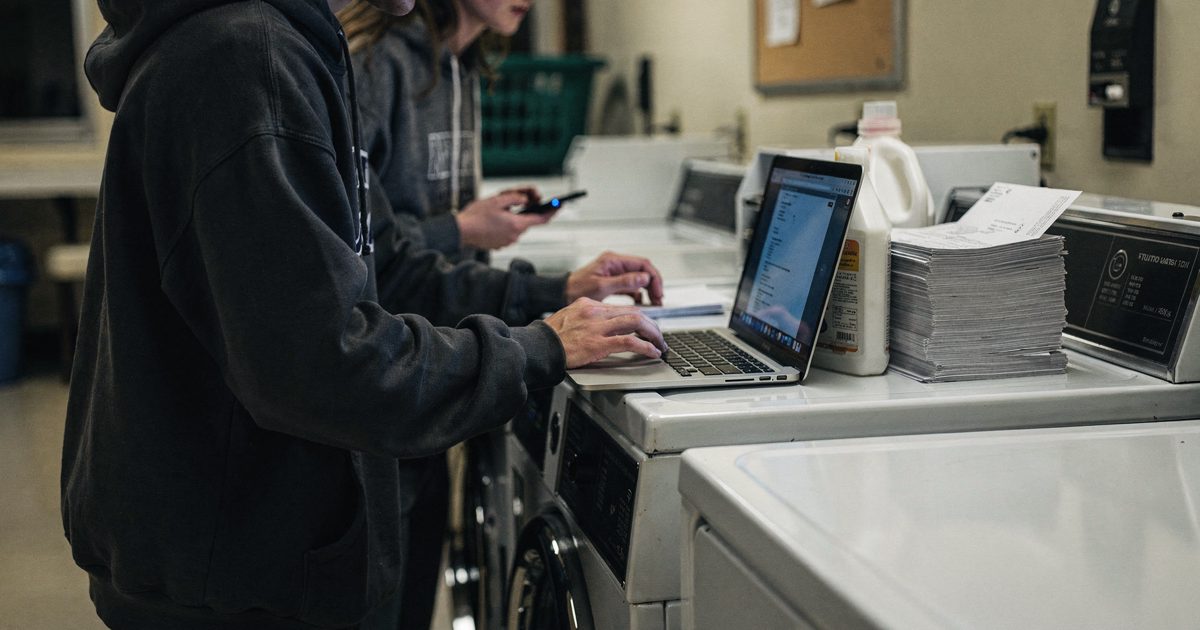 Late-evening dorm laundry room lit by overhead fluorescents, two part-cropped students in hoodies hunched over a laptop balanced on a running dryer, a stack of.