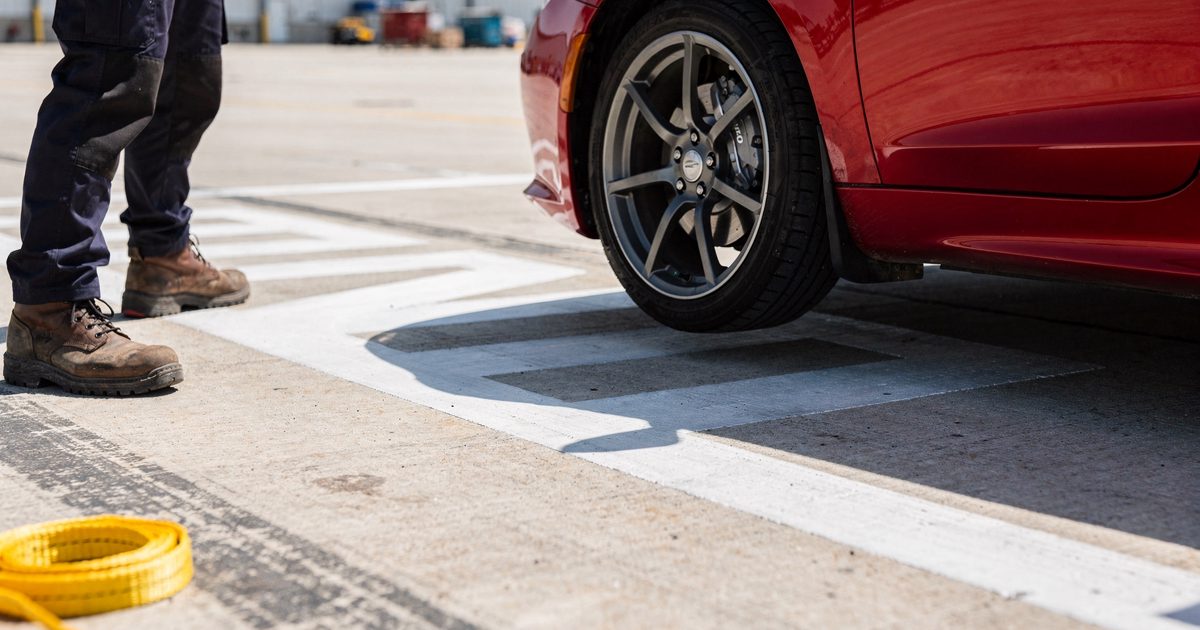 Low close detail in crisp, bright mid-morning sun on a concrete airfield test strip, the front wheel of a red sports car hanging a hand's width above painted.