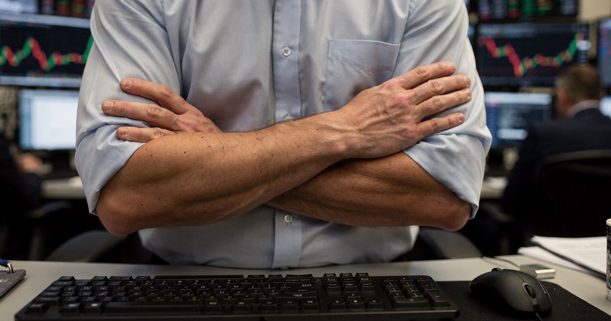 Close detail on a New York trading desk in bright early-morning light, a trader's shirtsleeved forearms crossed tightly over his chest above a keyboard while a.