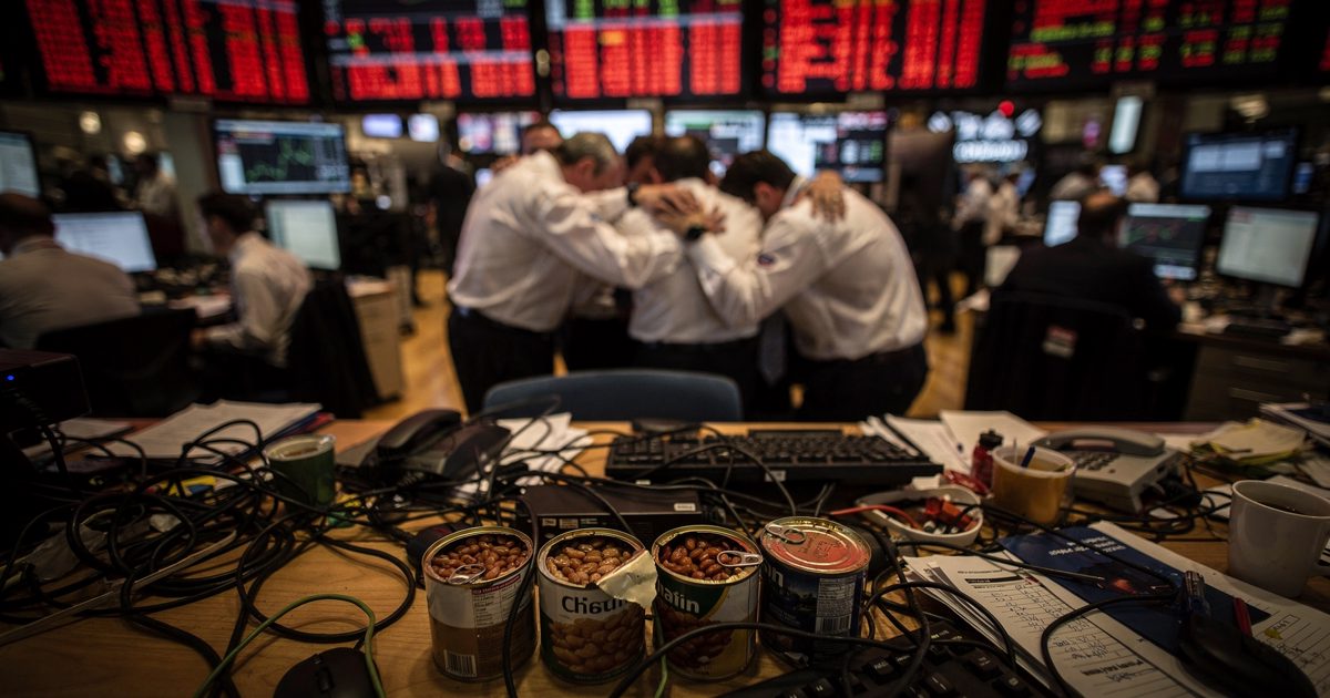 High-angle shot from the mezzanine of a crowded Manhattan trading floor in the harsh red glow of overhead ticker boards at dusk, a group of traders in white.