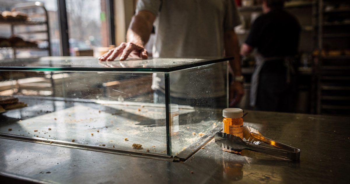 inside a neighborhood bakery in hard Sunday morning sidelight, a cropped customer from the chest down standing at an empty glass pastry case with one hand on.