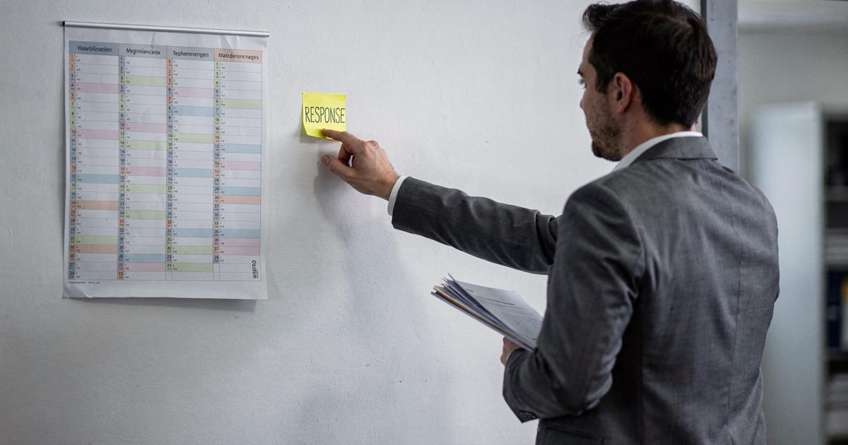 Medium shot in cool grey Geneva office light of a wall-mounted twelve-month year planner, a man in a grey suit seen from behind pressing a yellow sticky note.