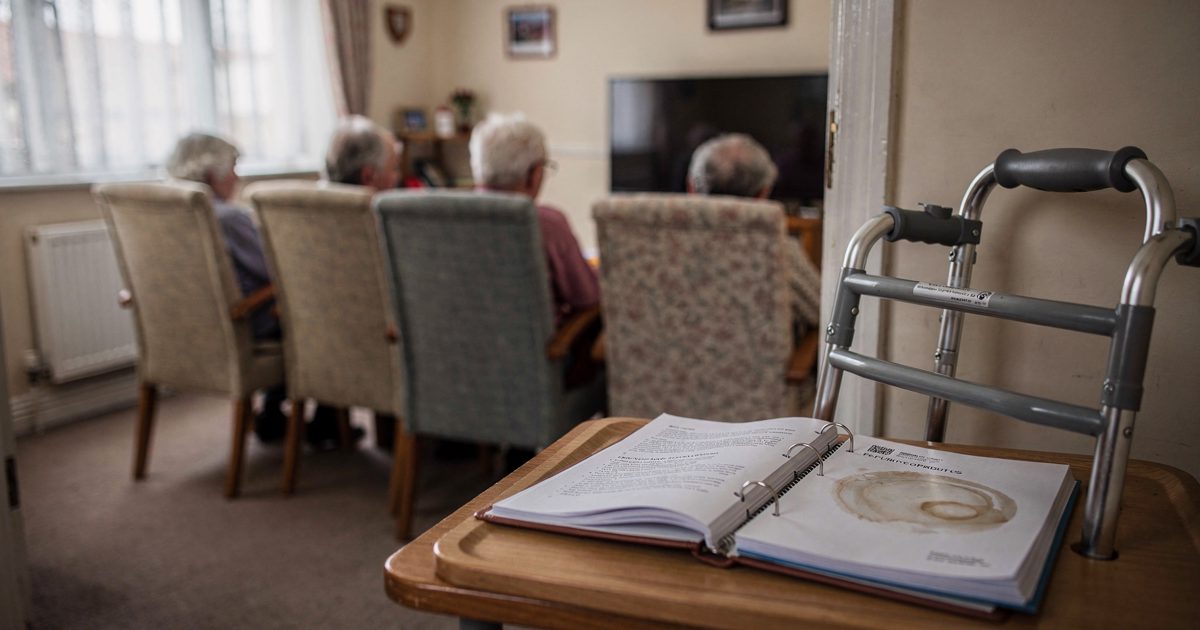 Medium shot from the doorway of a care home day room in soft morning light through net curtains, residents seen from behind in a row of high-backed chairs.