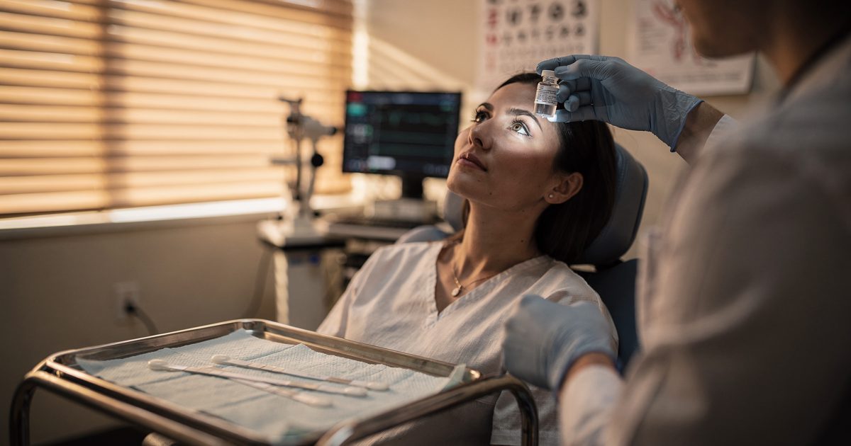 Warm early-evening light through the blinds of a hospital eye clinic as a patient framed from the jaw down tips her head back and a clinician's gloved hand.