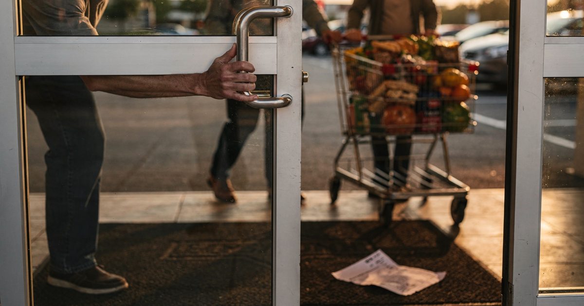 Close detail in warm late-afternoon light at a suburban grocery entrance: one shopper's hand braced against the glass door while a stranger pushes a loaded.