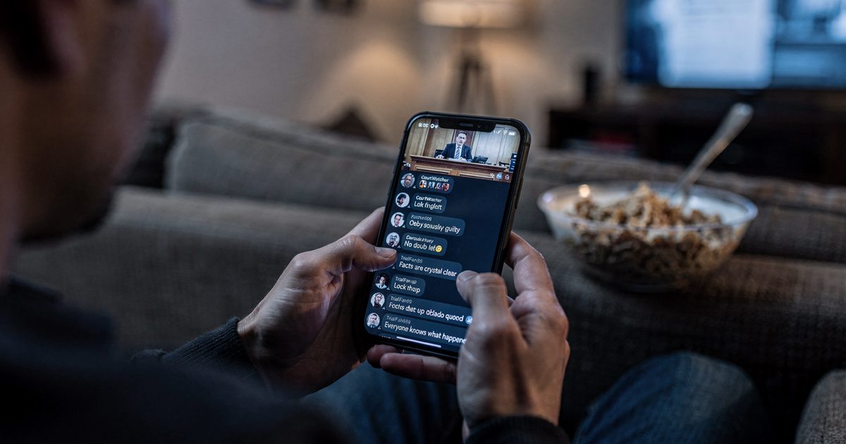 Close-up from behind of a viewer's hands cradling a phone at 11pm in a dim living room, courtroom livestream chat scrolling on the screen, a half-eaten bowl of.