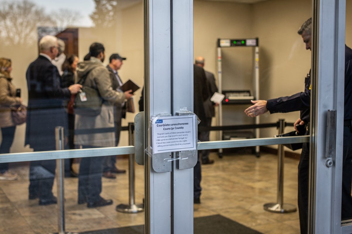 Wide daylight shot through the glass doors of a county courthouse lobby, a line of part-cropped figures filing past a metal detector in flat morning light, a.