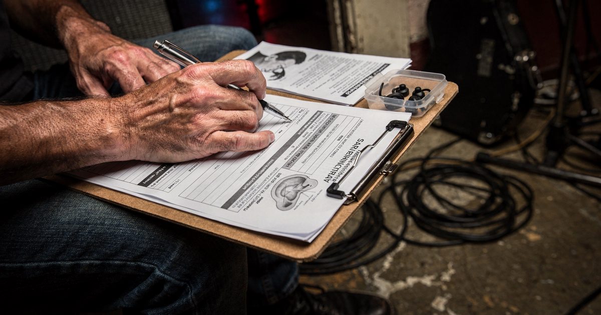 Close-up of a guitarist's calloused hands resting on a clipboard of insurance paperwork on his knees, backstage in a small Austin club under bare work lights.
