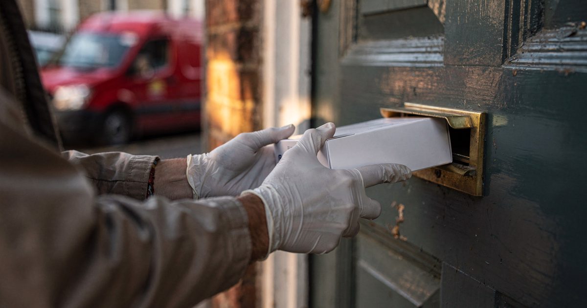 Close-up in low morning sun of gloved hands trying to angle a small white medical sample box through the brass mail slot of a terraced house's front door, a.