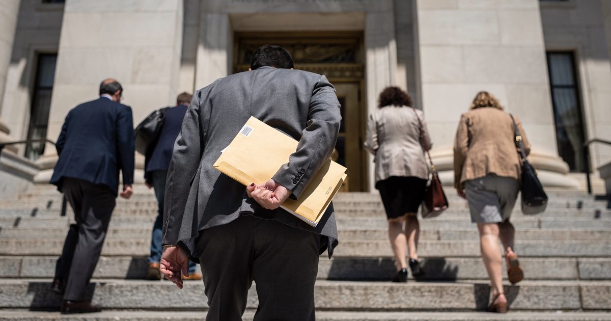 Harsh noon sun on the granite steps of a Massachusetts courthouse as several people seen only from the back climb toward the entrance, a sealed manila envelope.