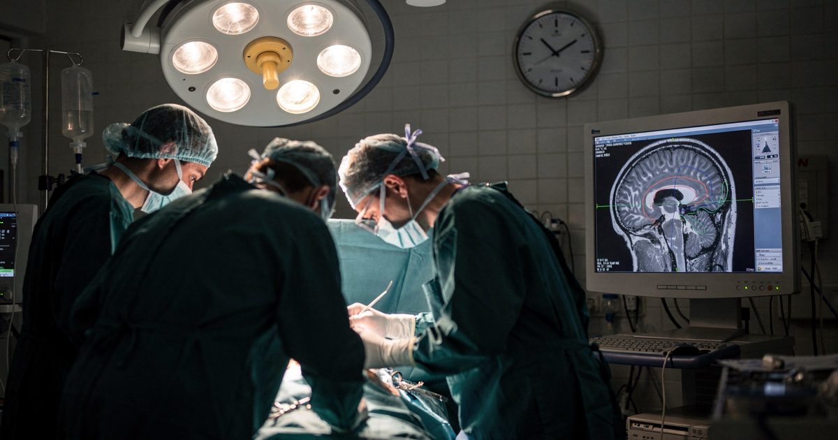Wide night-time view inside a working neurosurgical theatre under a bright overhead lamp array, gowned and masked figures bent over the table while a large.