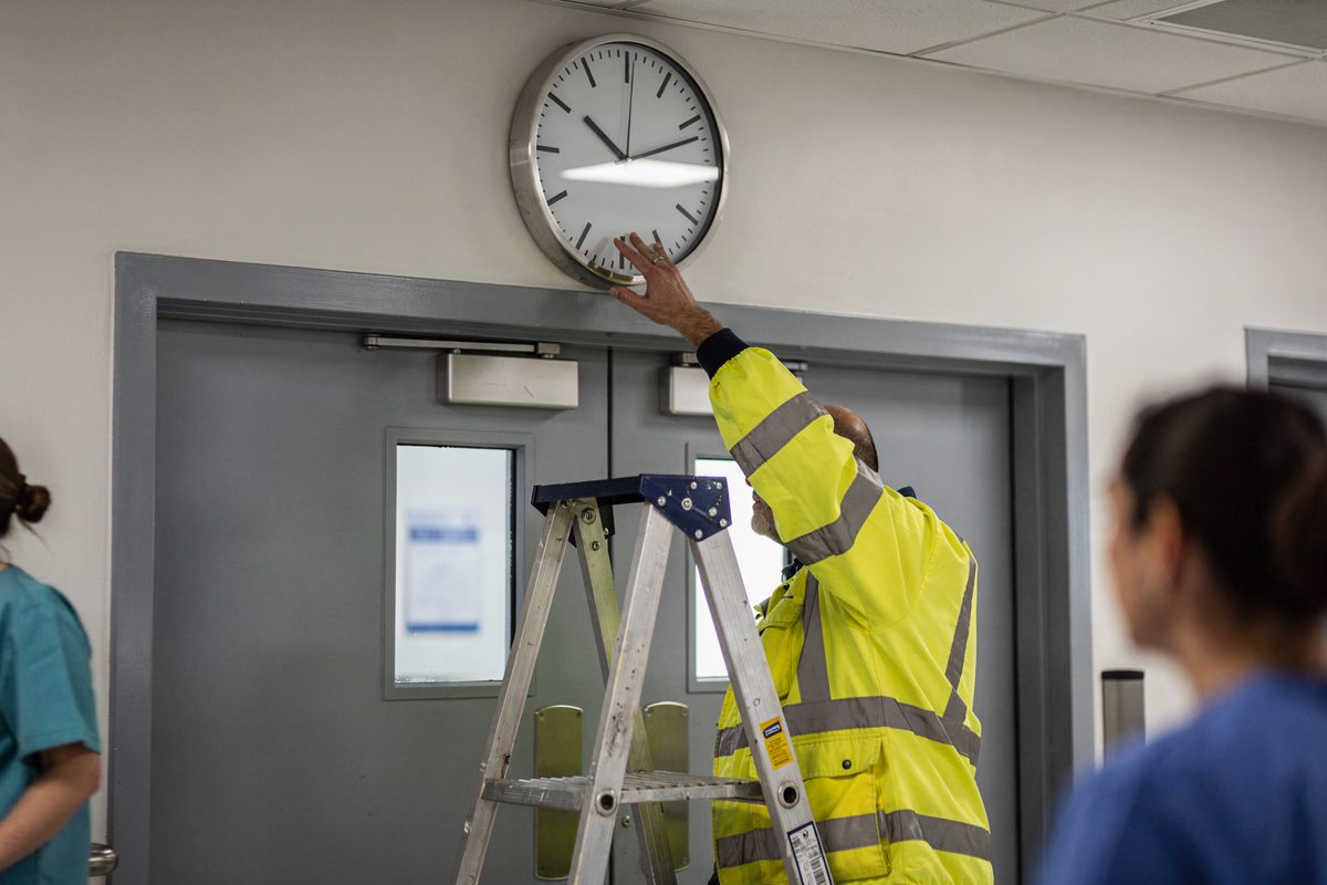 Low-angle mid-morning shot in a hospital corridor of a plain wall clock mounted above double theatre doors, a stepladder set beneath it and an inspector's.