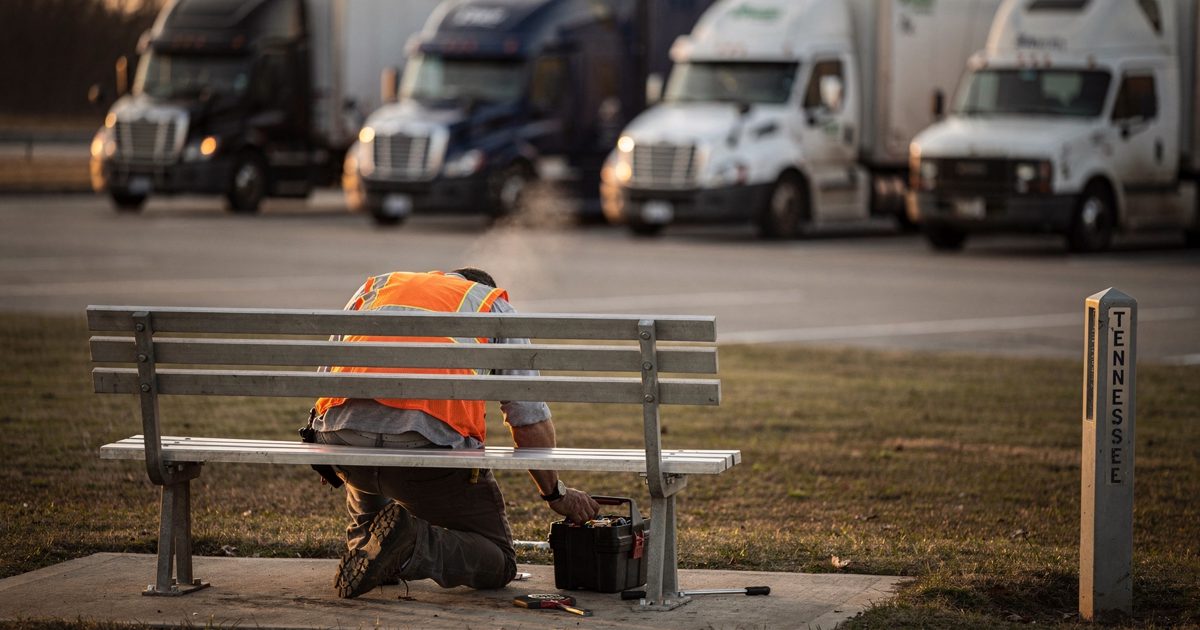 Wide shot across an interstate rest area in low golden dusk light with trucks idling in the background, a highway maintenance worker seen from behind in an.