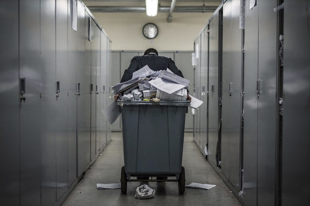 Wide shot from behind of a worker pushing a wheeled bin of loose paper down a windowless basement corridor lined with grey filing cabinets, fluorescent tubes.