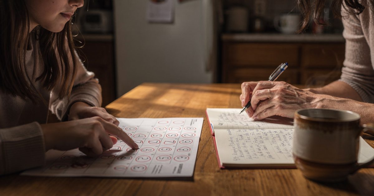Late-afternoon kitchen light on a wooden table as a younger hand points at red-circled dates on a paper calendar while an older pair of hands hovers a.