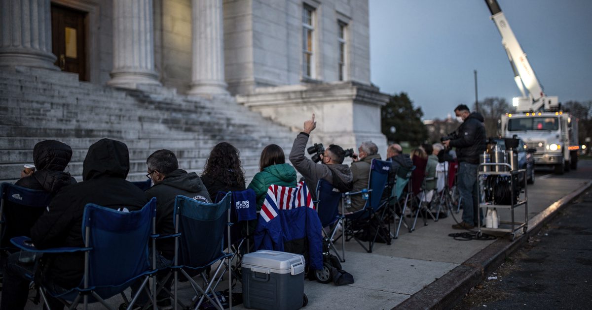 Pre-dawn outside a granite county courthouse, a queue of spectators seen from behind settling into folding camp chairs, a satellite truck's mast rising against.