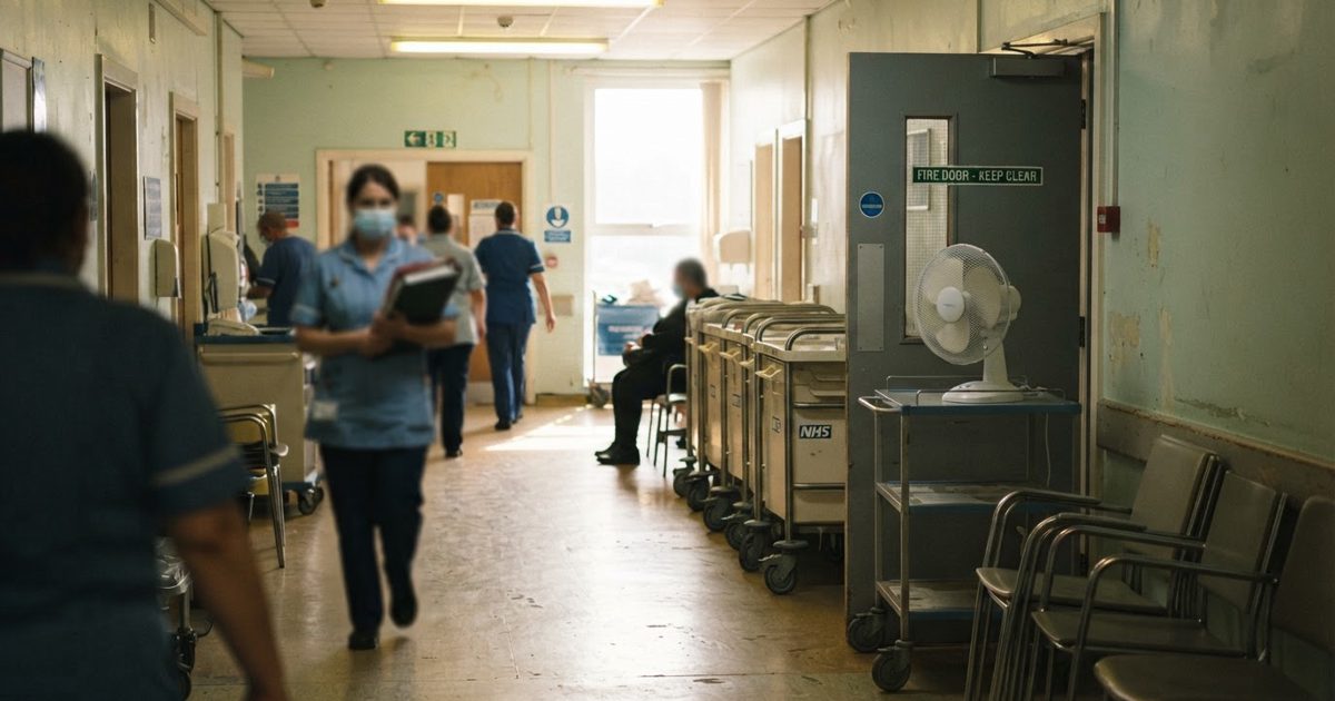 Documentary-style news photograph of a hospital corridor in a British NHS building on a bright summer afternoon: empty wheeled trolleys and stacked chairs.