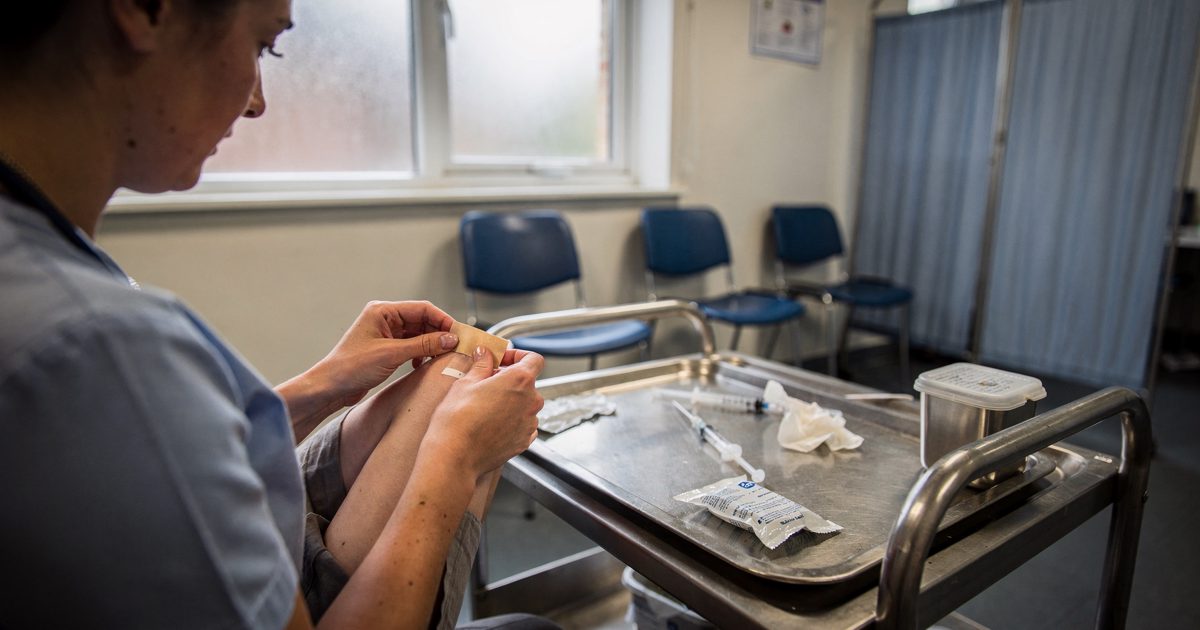 Pale morning light through a high window in an NHS walk-in centre as a vaccinator seen from behind at the shoulder applies a small plaster to a patient's upper.
