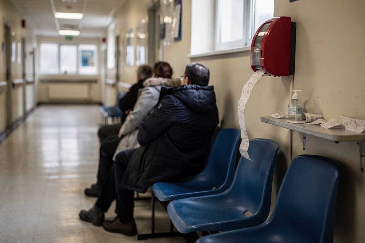 Wide shot down a long community health centre corridor on a bright Saturday morning with hard winter light bouncing off the lino, three people in winter coats.