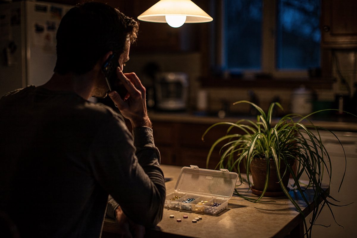 Mid-shot at dusk in a small home kitchen lit by one overhead pendant, a person seen from behind at the table holding a phone to their ear on speaker, a.