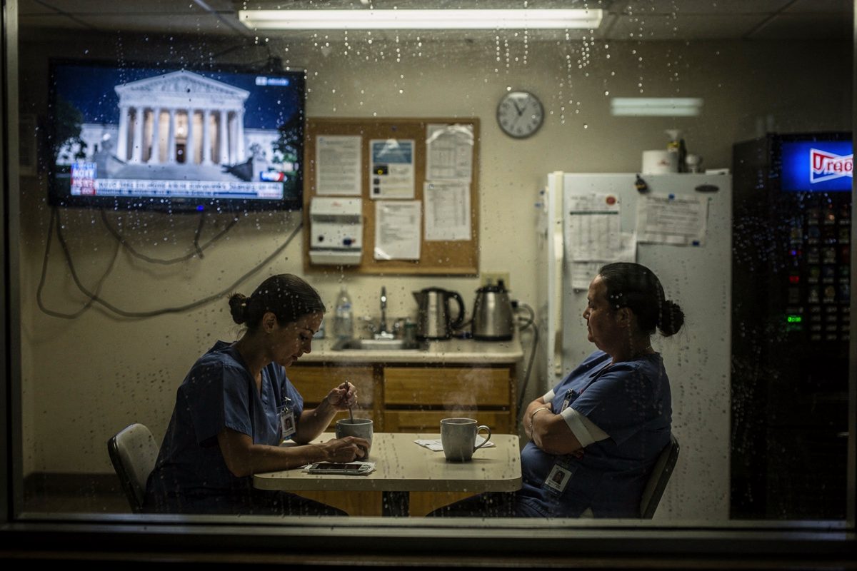 Medium shot at night through a rain-streaked window of a small hospital break room, two night-shift nurses cropped at the chin sitting with tea while a silent.