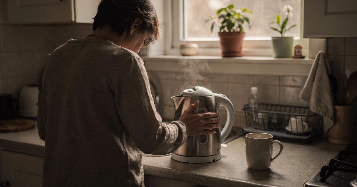 Morning light through a small kitchen window, a person seen from behind standing at the counter with one hand on an electric kettle and an unused second mug by.
