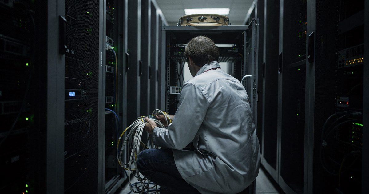 Night shot down a narrow server hall aisle lit only by rack indicator lights, a technician in an anti-static smock crouched at an open cabinet with cables in.
