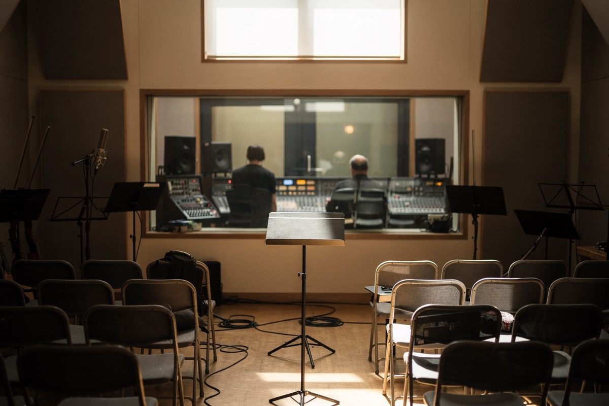 Wide midday shot of a small recording studio live room with daylight from a high window, rows of unused chairs where a string section would sit, two figures.