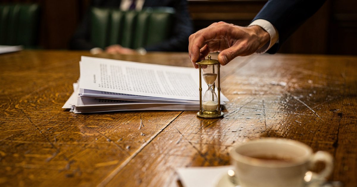Close shot in slanted late-afternoon light on a scarred oak table in a Westminster committee room, a hand in a navy sleeve setting a small brass hourglass.