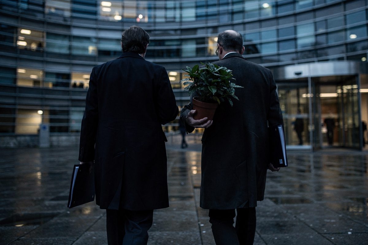 Medium shot in thin early-morning blue on the wet plaza before a curved glass EU office block, two officials seen only from behind walking toward the doors.
