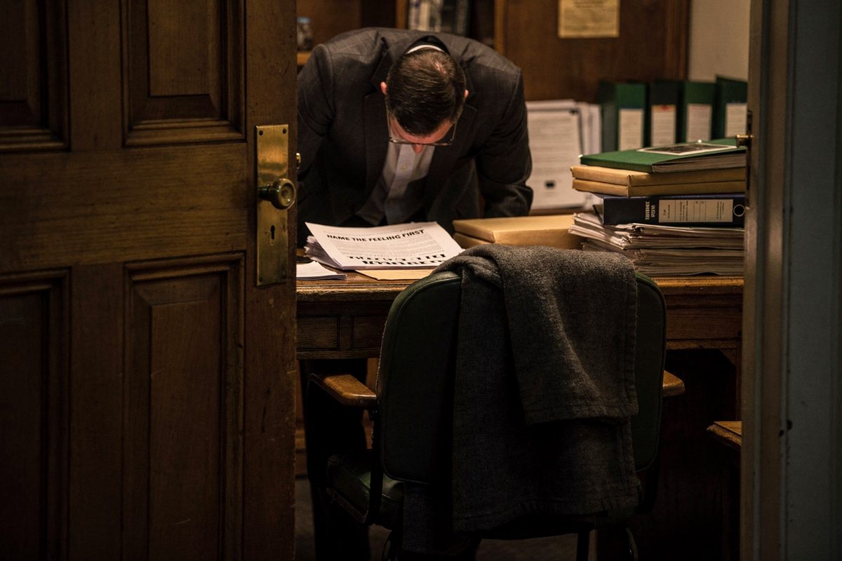 Night view through a half-open oak door of a Commons clerks' room, a person leaning over a desk with head bowed so only the crown of hair shows, lamplight on.