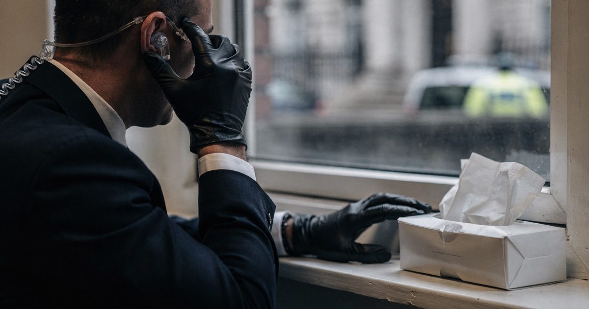 Close-up in soft morning light of a close-protection officer's gloved hands adjusting a radio earpiece beside an open box of tissues on a Whitehall windowsill.