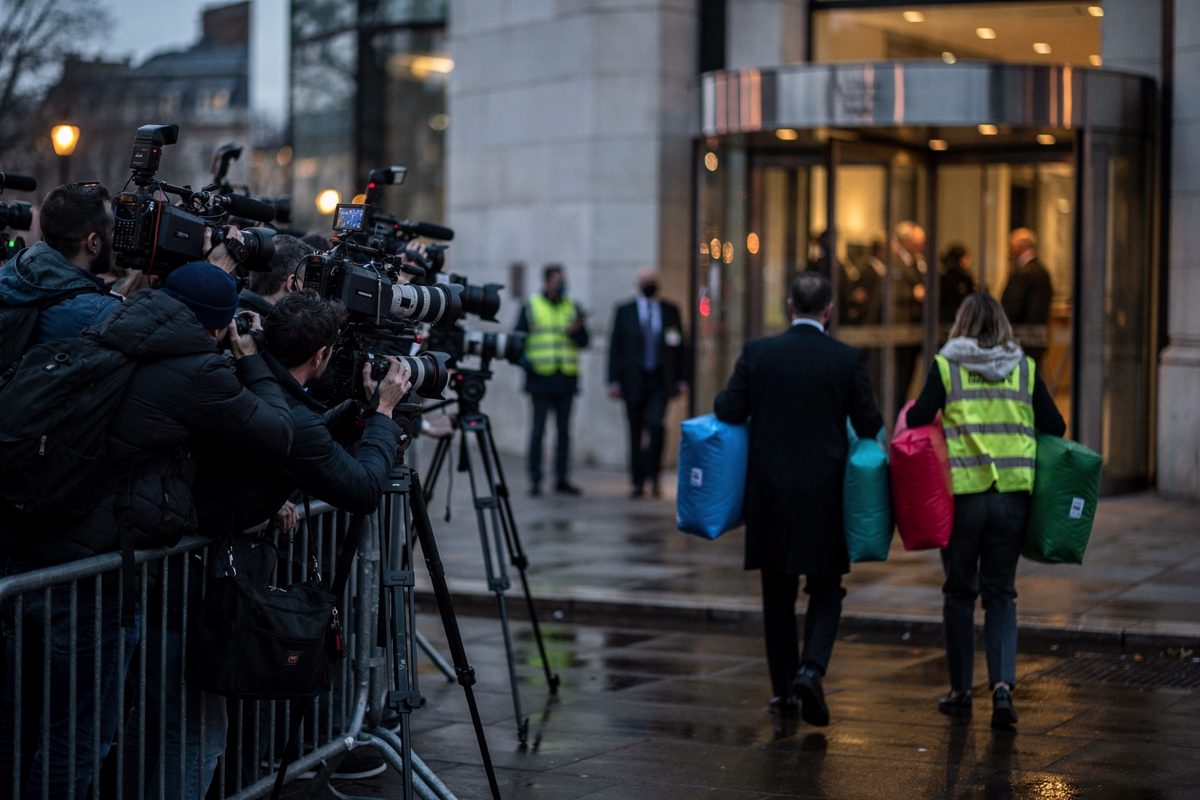 Wide shot at dusk outside a government building in Westminster, photographers with long lenses crowding a barrier while two staffers, seen from behind, carry.