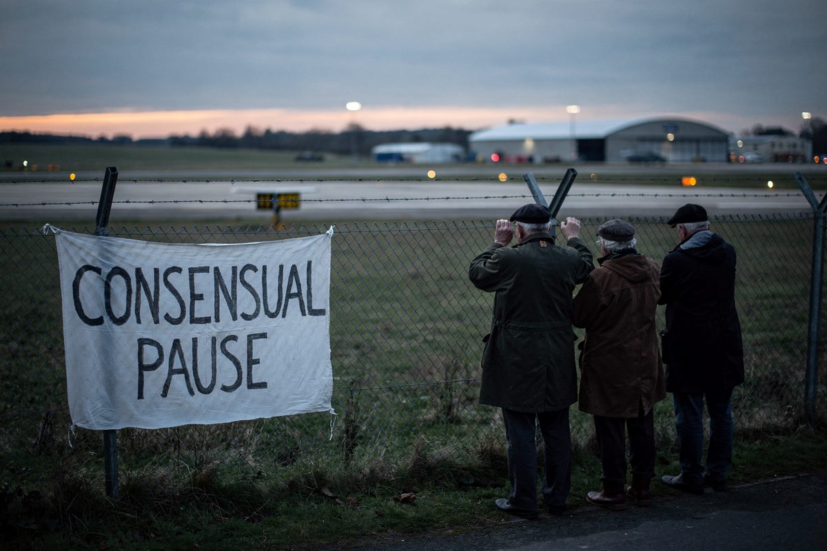 Wide shot along a chain-link perimeter fence at an airfield in Gloucestershire at dusk, flat farmland and a long empty runway beyond, three villagers in waxed.