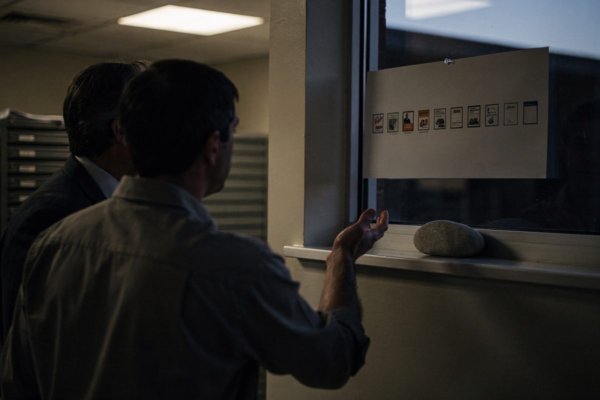 Over-the-shoulder dusk light in a state office annex, two people cropped at the neck marking a wall calendar with a row of fourteen dated stickers, a smooth.