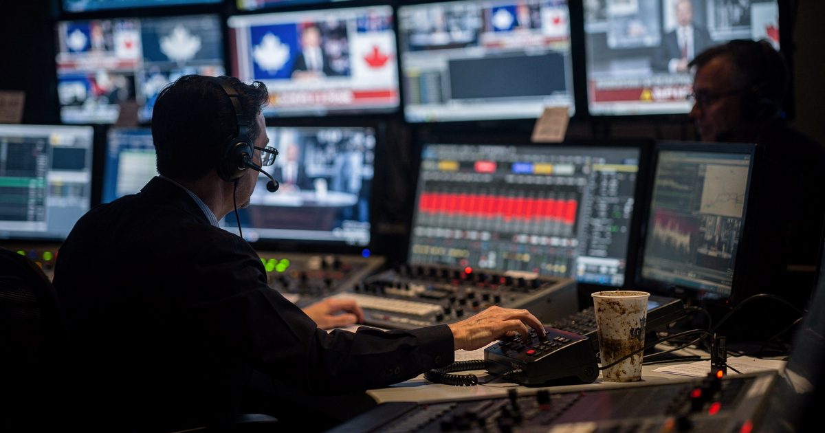 Wide night shot of a television network control room, a bank of glowing monitors washing two headset-wearing producers in blue light as one reaches across the.