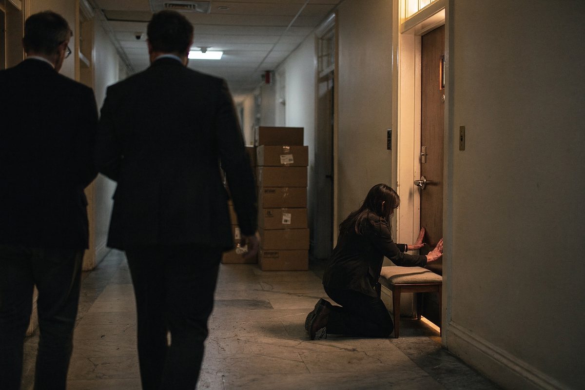 Figures seen from behind in a narrow Capitol side hallway at dusk, one person setting a small floor cushion against a wooden doorframe while warm light from a.