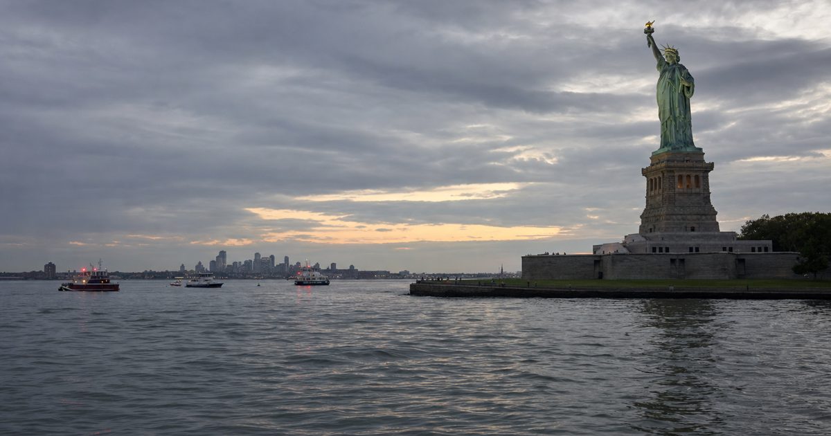 Respectful wide news photograph of New York Harbor near Liberty Island at dusk, calm water in the foreground, emergency boats visible at a great distance, no.