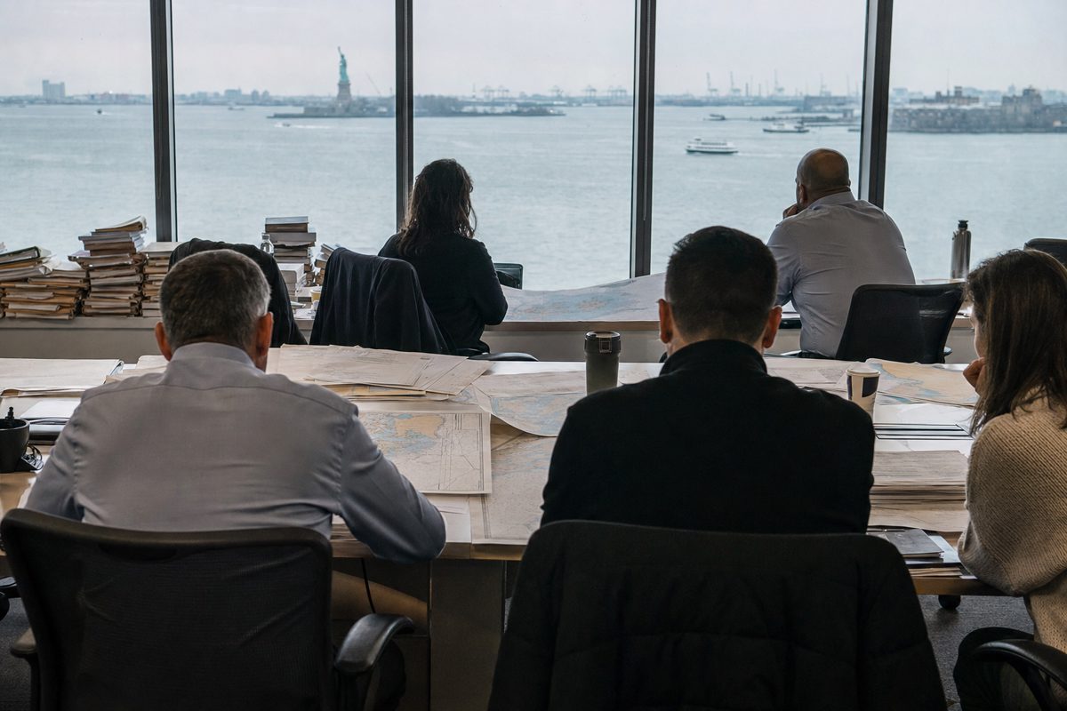 News photograph inside a generic municipal conference room overlooking New York Harbor, anonymous officials seen from behind studying nautical charts and.