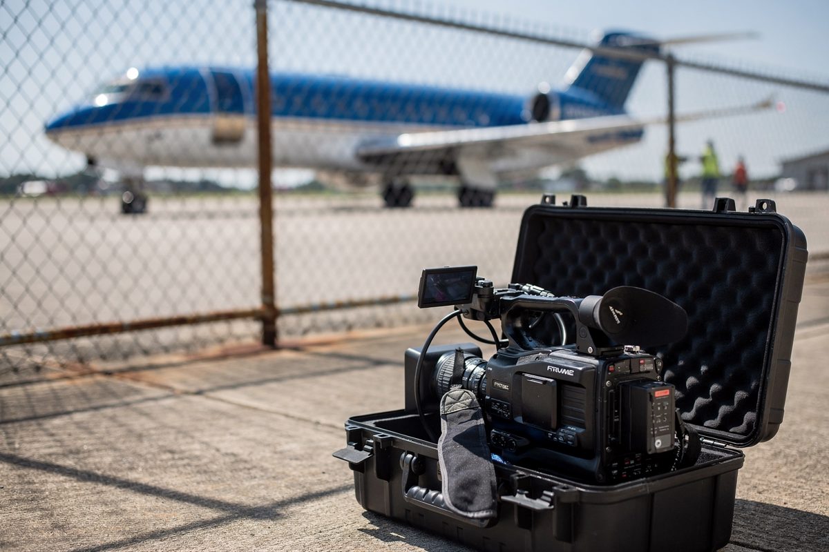 Close detail in hard late-morning sun at the edge of a regional airport apron in Tennessee, a broadcast camera in a hard-shell case left standing on the.
