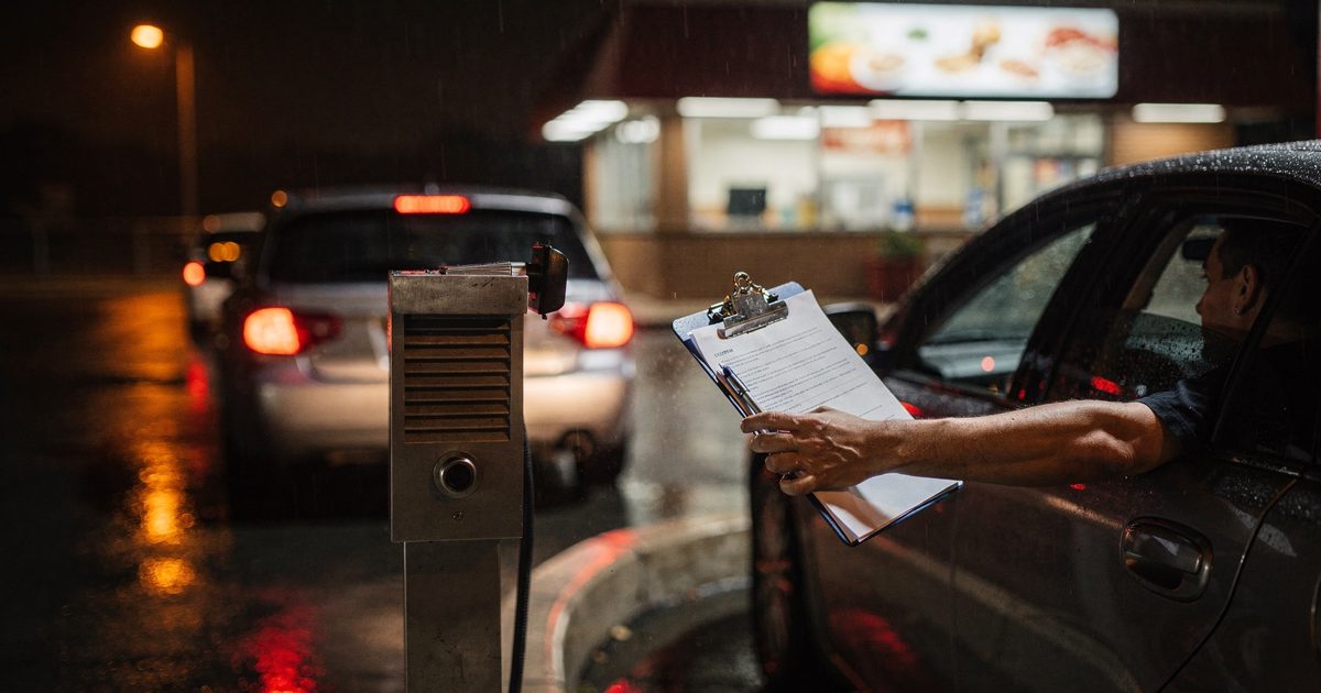 Night shot from behind a car at a fast-food drive-thru speaker, headlights flaring off wet asphalt, a driver's arm extended out the window holding a clipboard.
