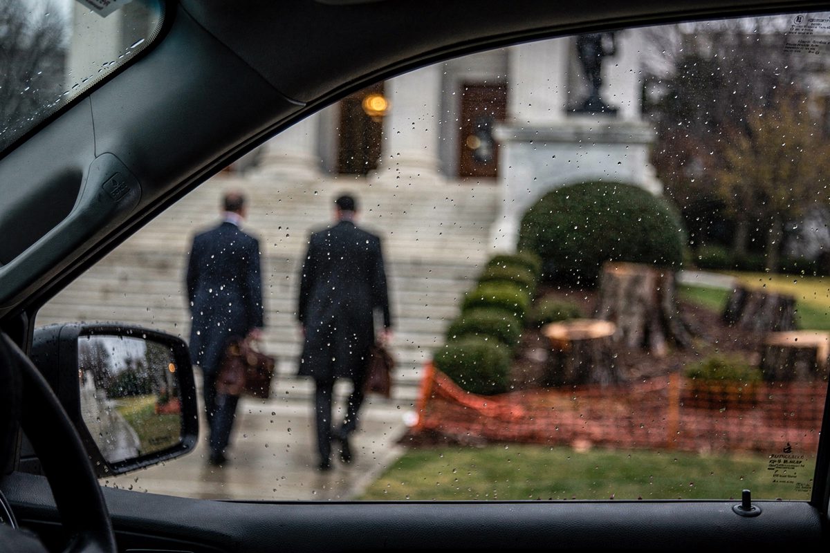 Medium shot through the rain-streaked passenger window of a parked black SUV outside a neoclassical Washington courthouse at midday, two lawyers in dark suits.