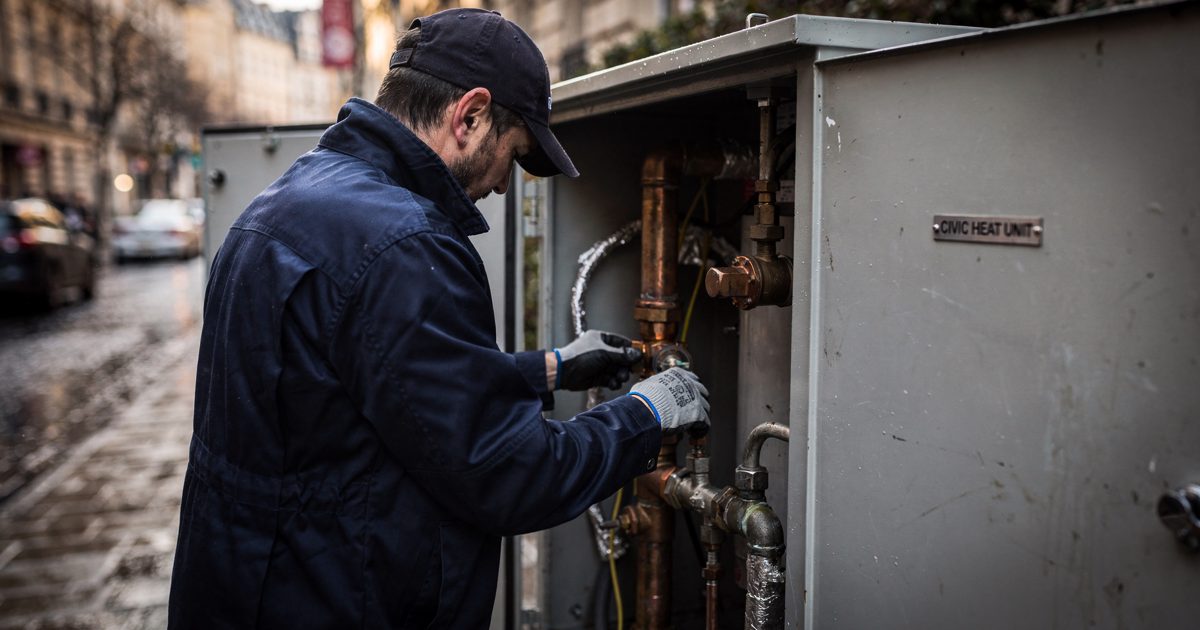 Close late-afternoon sunlight on a grey metal street cabinet on a cobbled European sidewalk as a technician in a navy work coat, head bowed so only a cap brim.