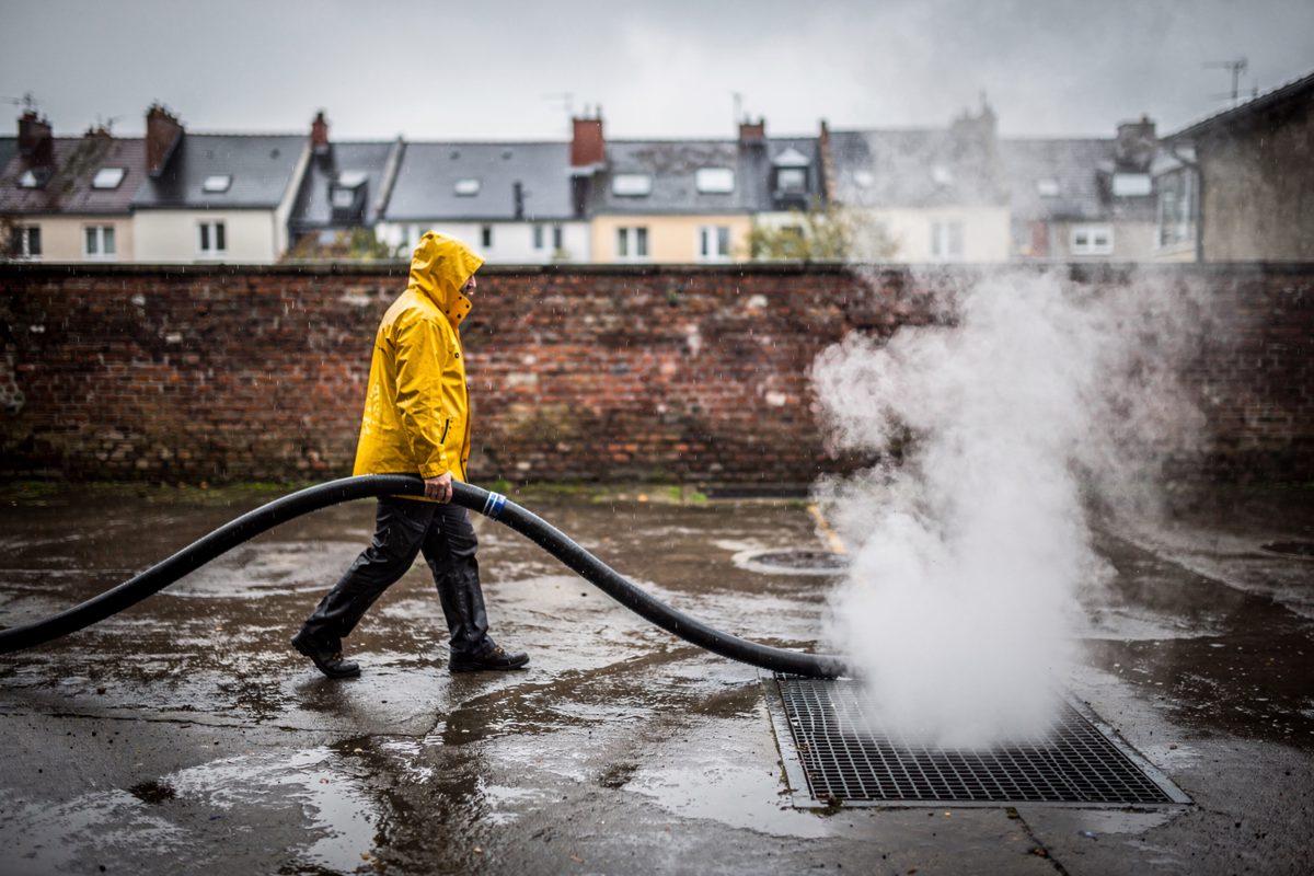 Wide rain-soaked midday across the yard of a brick district-heating plant as a worker in a yellow waterproof, cropped at the shoulders, drags a heavy hose.