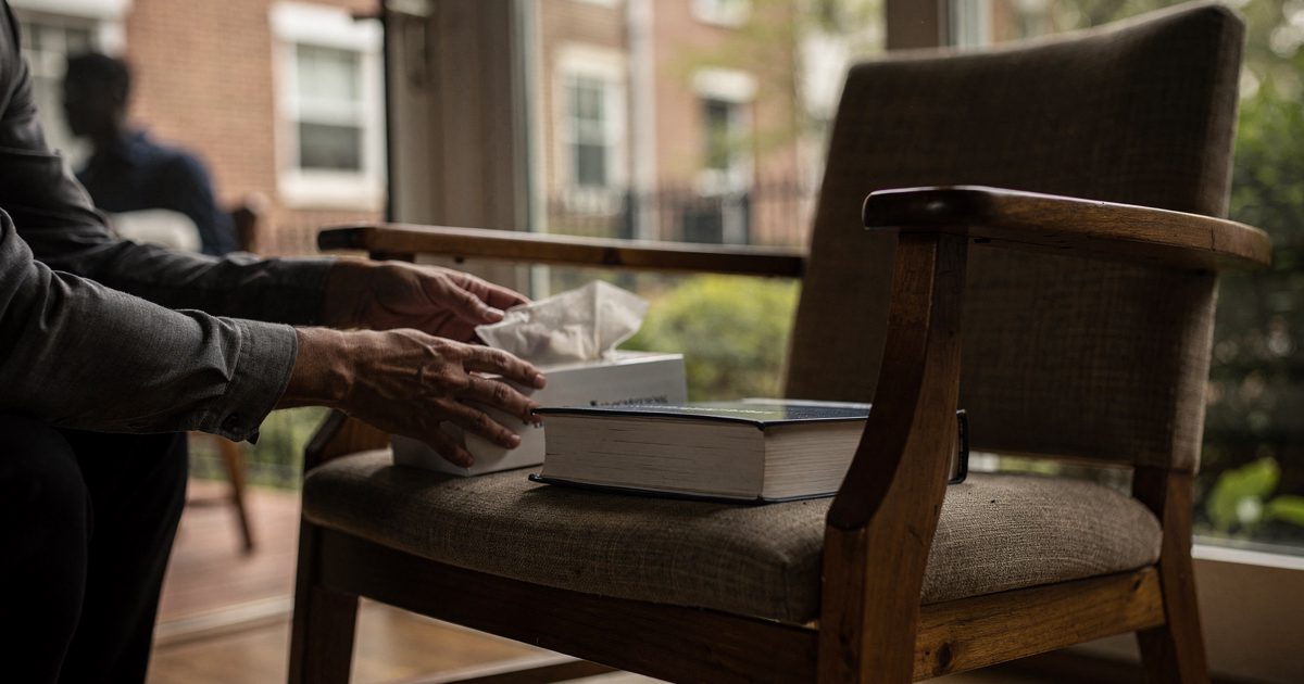 Close detail in soft late-morning light through a glass sunroom in a Washington rowhouse, a facilitator's hands, cropped at the wrists, setting a box of.