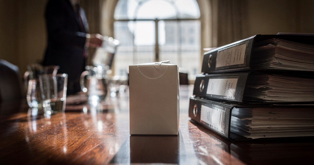 Close detail in hard morning sunlight through a tall arched window of an unopened box of tissues standing upright on a long polished mahogany boardroom table.